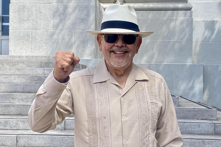 Image of Carlos Muñoz smiling at the camera, raising his fist, wearing sunglasses and a panama hat