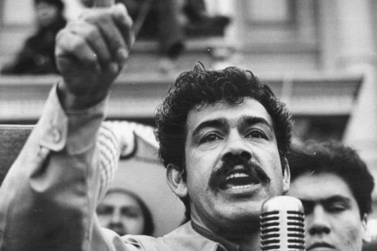Activist Corky Gonzalez raising his hand and speaking into a microphone in front of a crowd