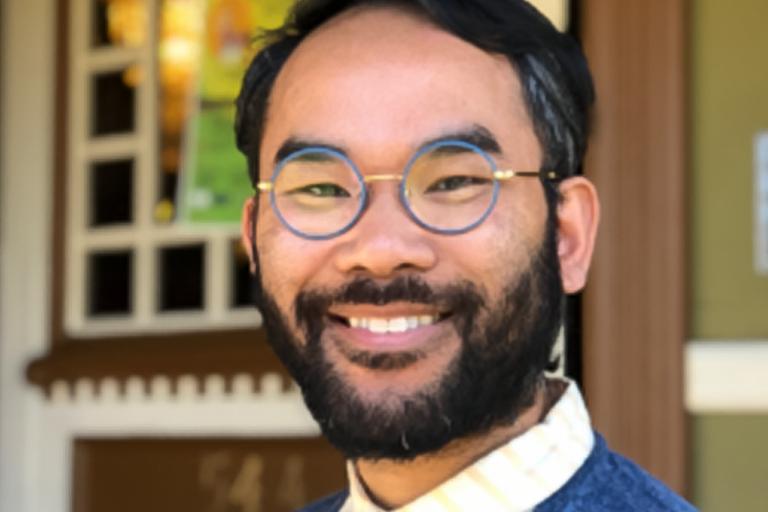 Photo of Elijah Chhum, smiling, wearing glasses and a sweater over a bright collared shirt in front of the facade of a building