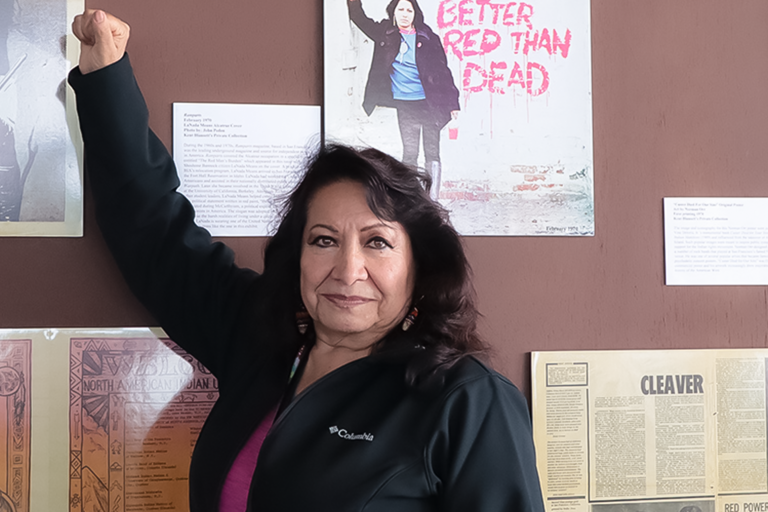 Body shot of LaNada WarJack standing her fist raised at a museum exhibit that features posters of indigenous movements