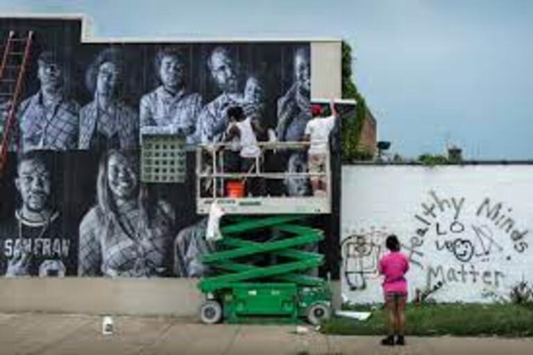 Woman stands in front of two individuals on a lift ladder posting a wallpaper mural of Black bay area community members
