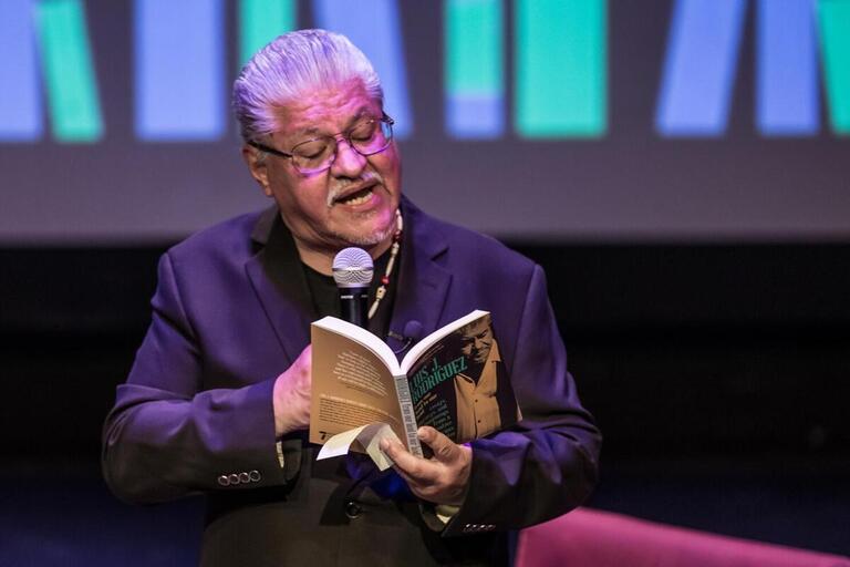 Luis Rodriguez reading from a book and speaking into a microphone, wearing a suit jacket and a beaded necklace