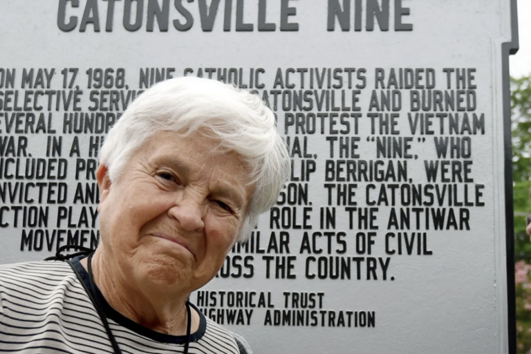 Margarita Mellville smiling in front of a placard for the Catsonville Nine