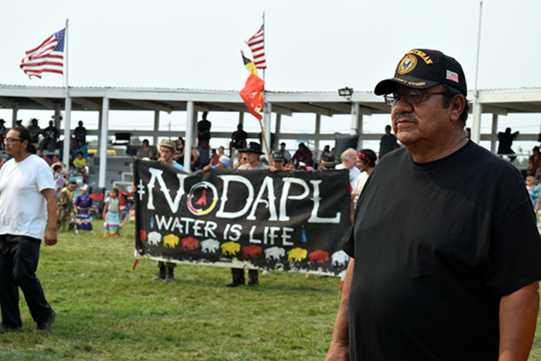 Man in the foreground. In the background is a group of people holding a sign that says "NoDAPL Water is Life". 