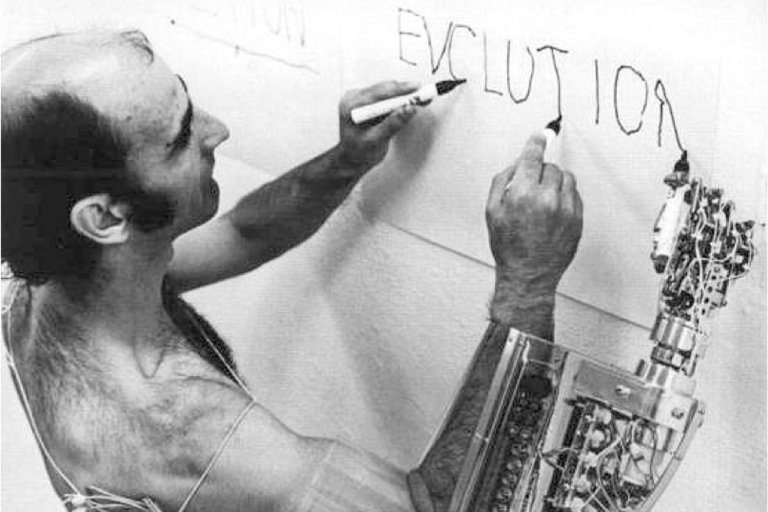 Black and white image of a man writing "Evolution" using both hands on a piece of paper on a wall using both hands, man is able to write with right hand due to assistive technology
